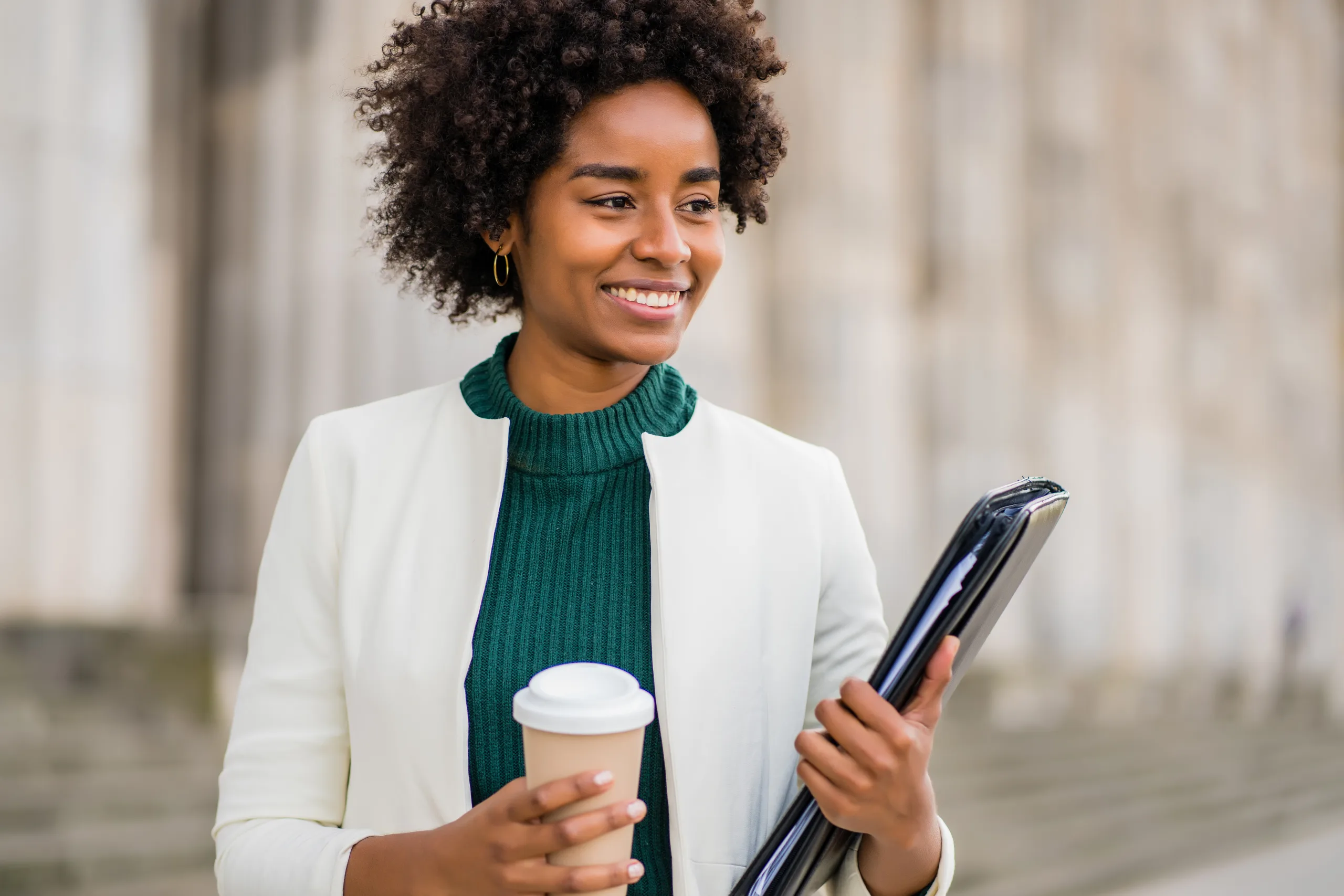 businesswoman smiling outdoors, holding a coffee cup and a folder, representing confidence and professionalism in a business environment.