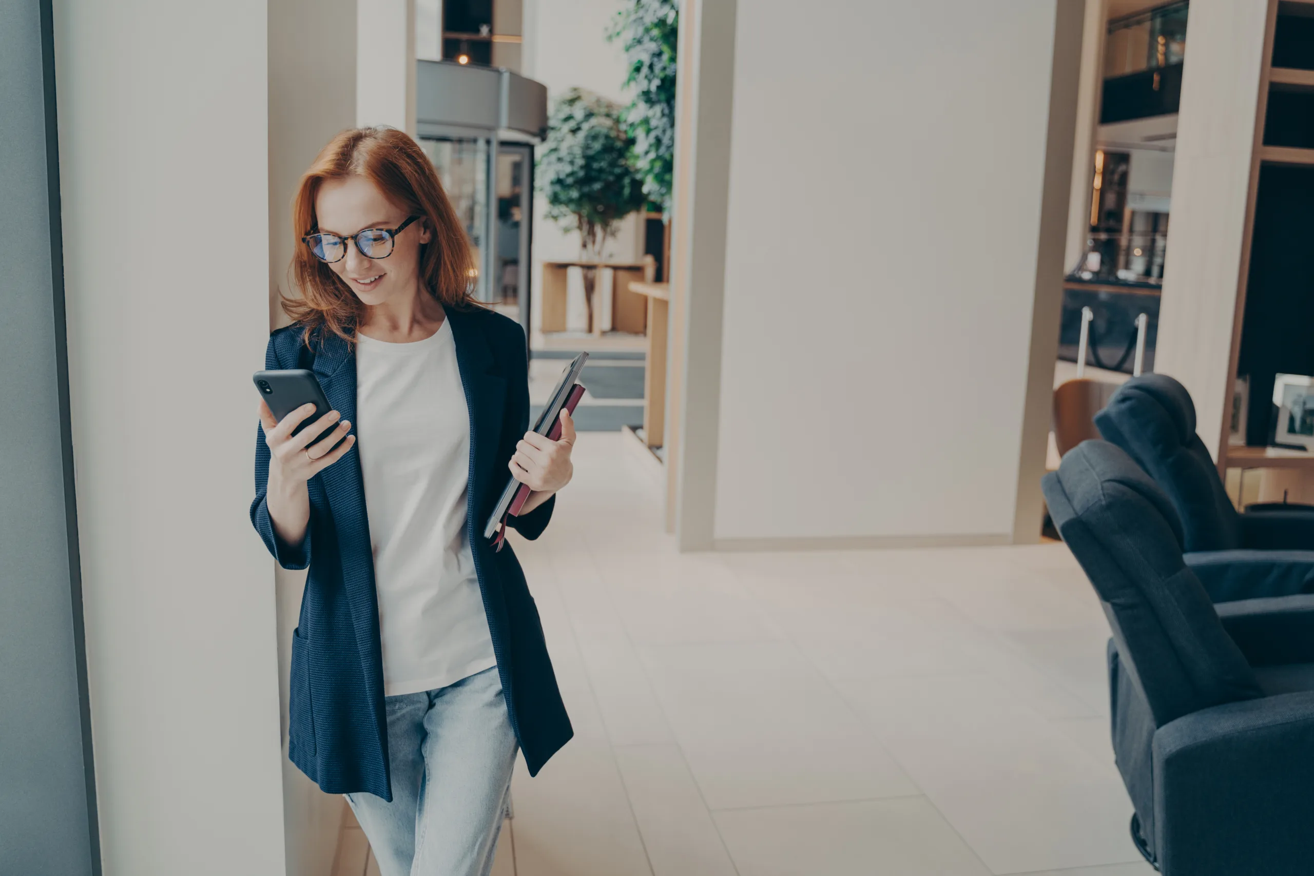 Smiling redhead female business consultant in a blazer, holding a smartphone and documents, standing in a modern office environment, reflecting Sanguine Strategic Advisors' mission to support business growth and innovation.
