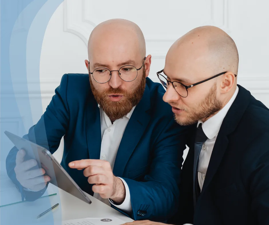 Bald men in business suits discussing strategies while reviewing a tablet, representing collaboration and problem-solving at Sanguine Strategic Advisors.