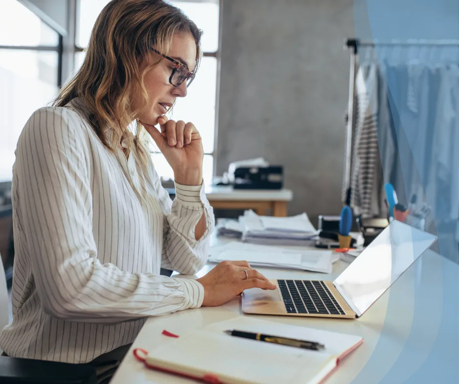 Woman working thoughtfully on a laptop in a modern office environment, surrounded by paperwork and office supplies, reflecting a professional atmosphere aligned with Sanguine Strategic Advisors' mission to support business growth and success.