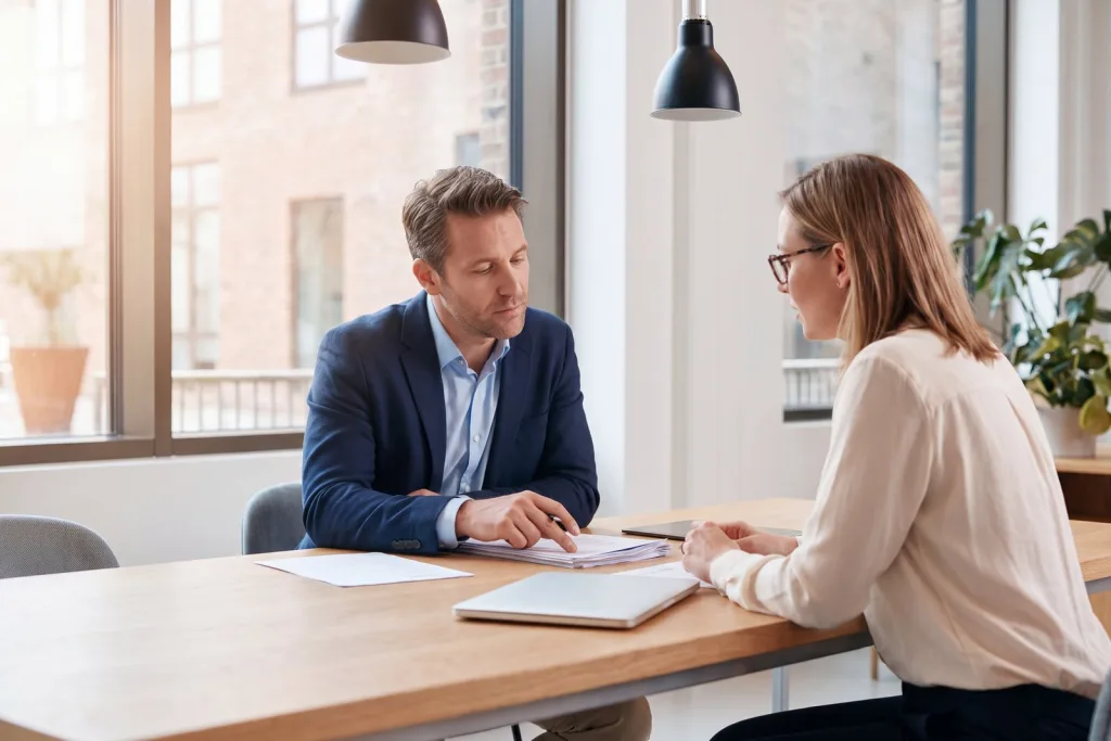 Two people in a quiet, focused conversation at a table, one hand resting thoughtfully on a printed document